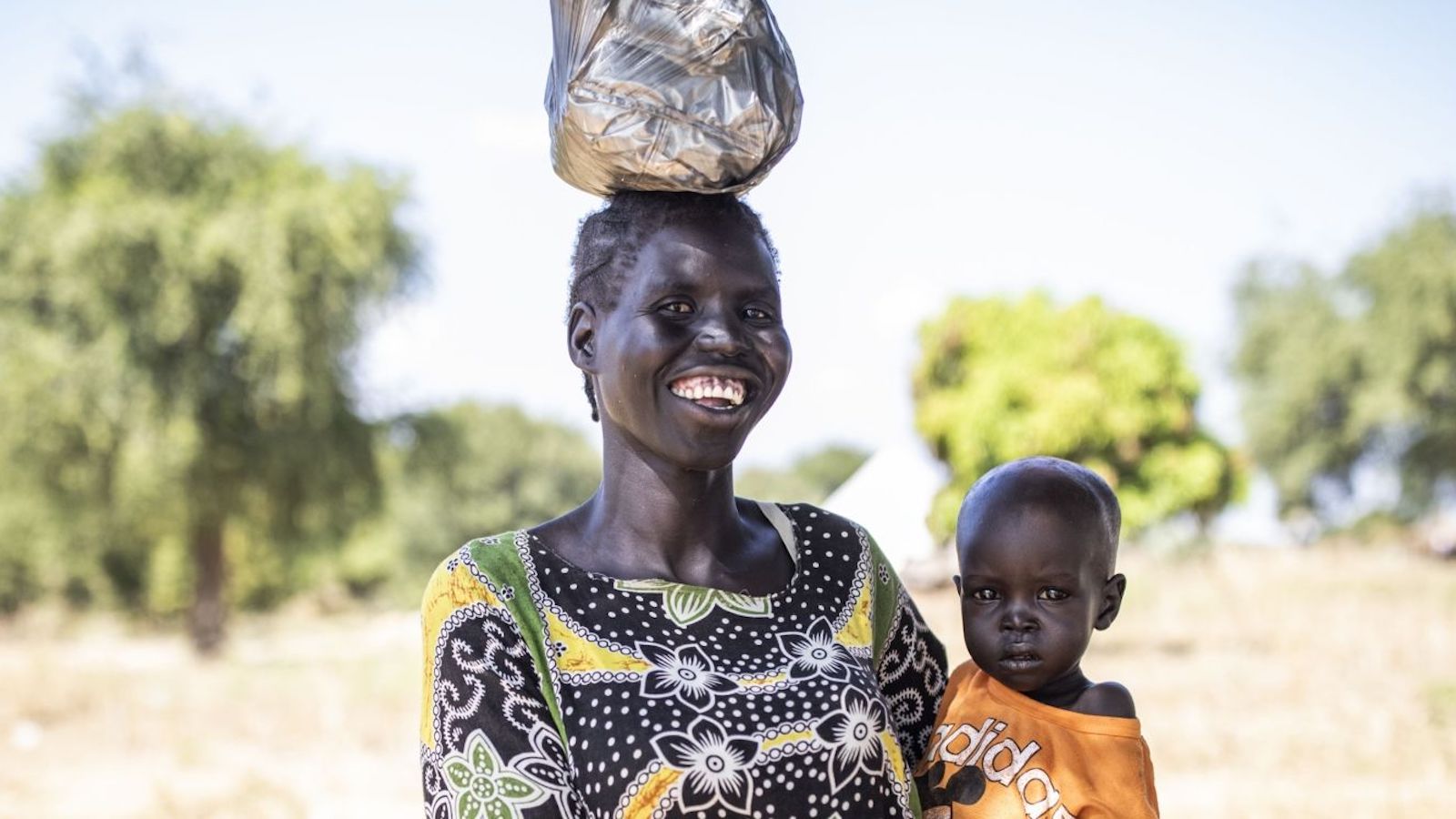 Nyibar holds infant son in her arms and balances bag on her head in South Sudan