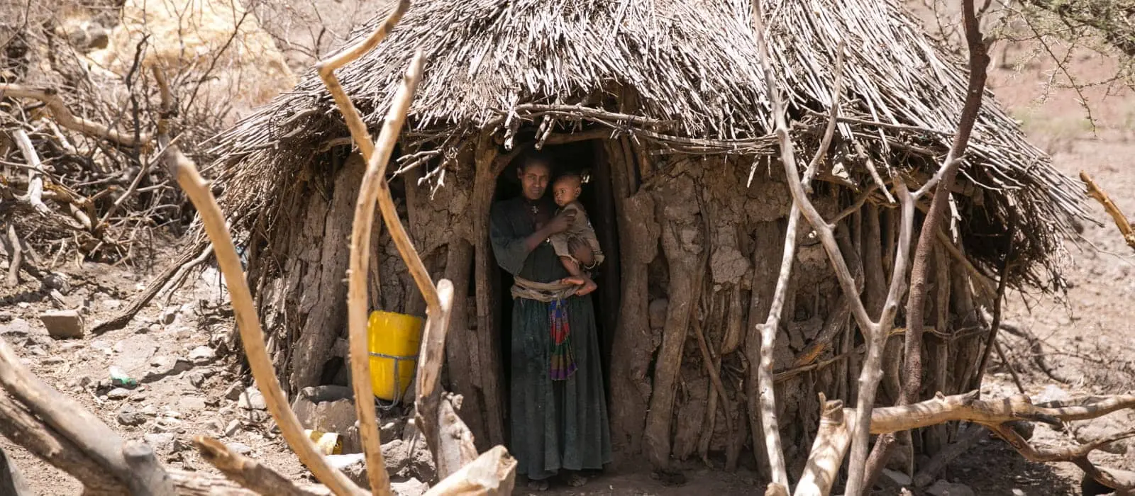 Woman and child stand in doorway of mud and thatch hut in barren Ethiopan landscape