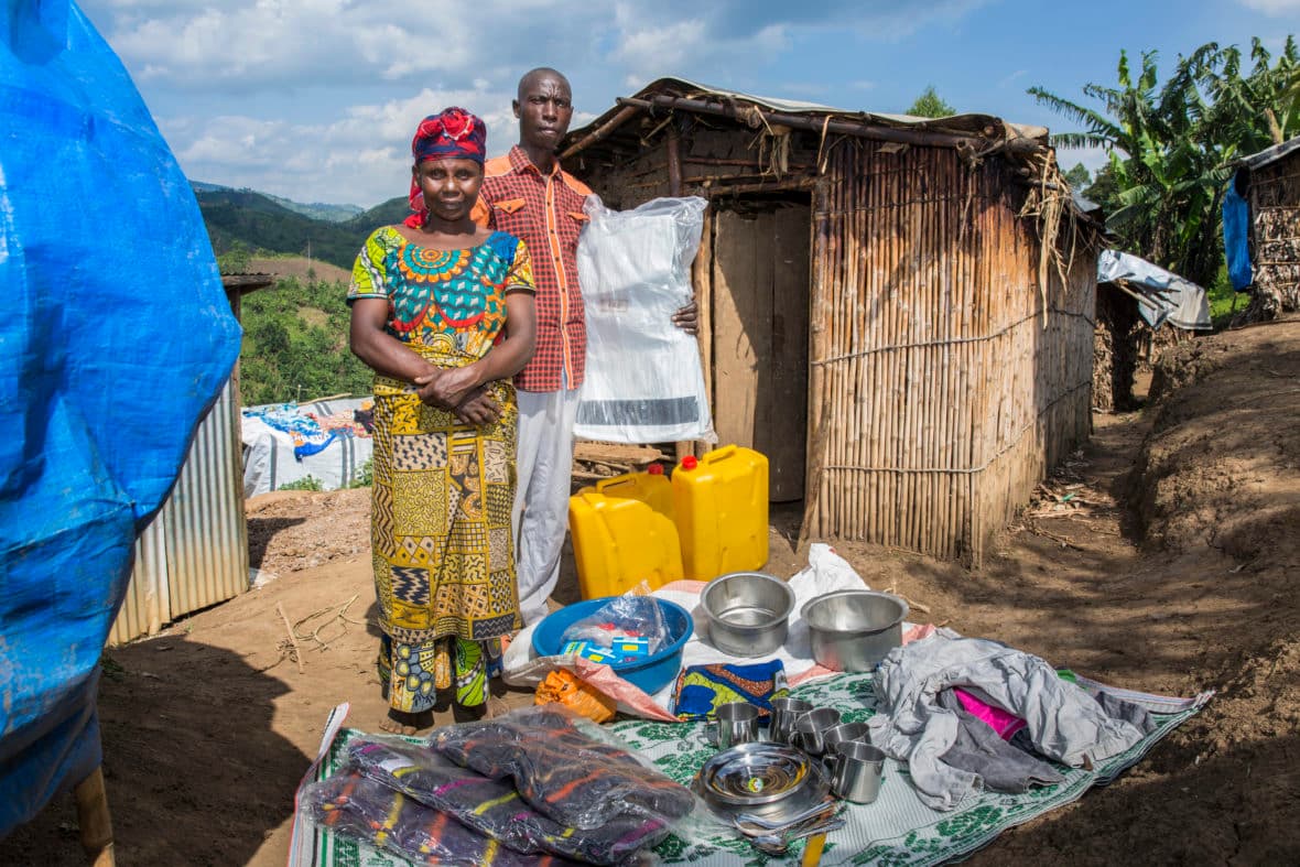 A couple standing outside a temporary shelter on a hillside with household supplies