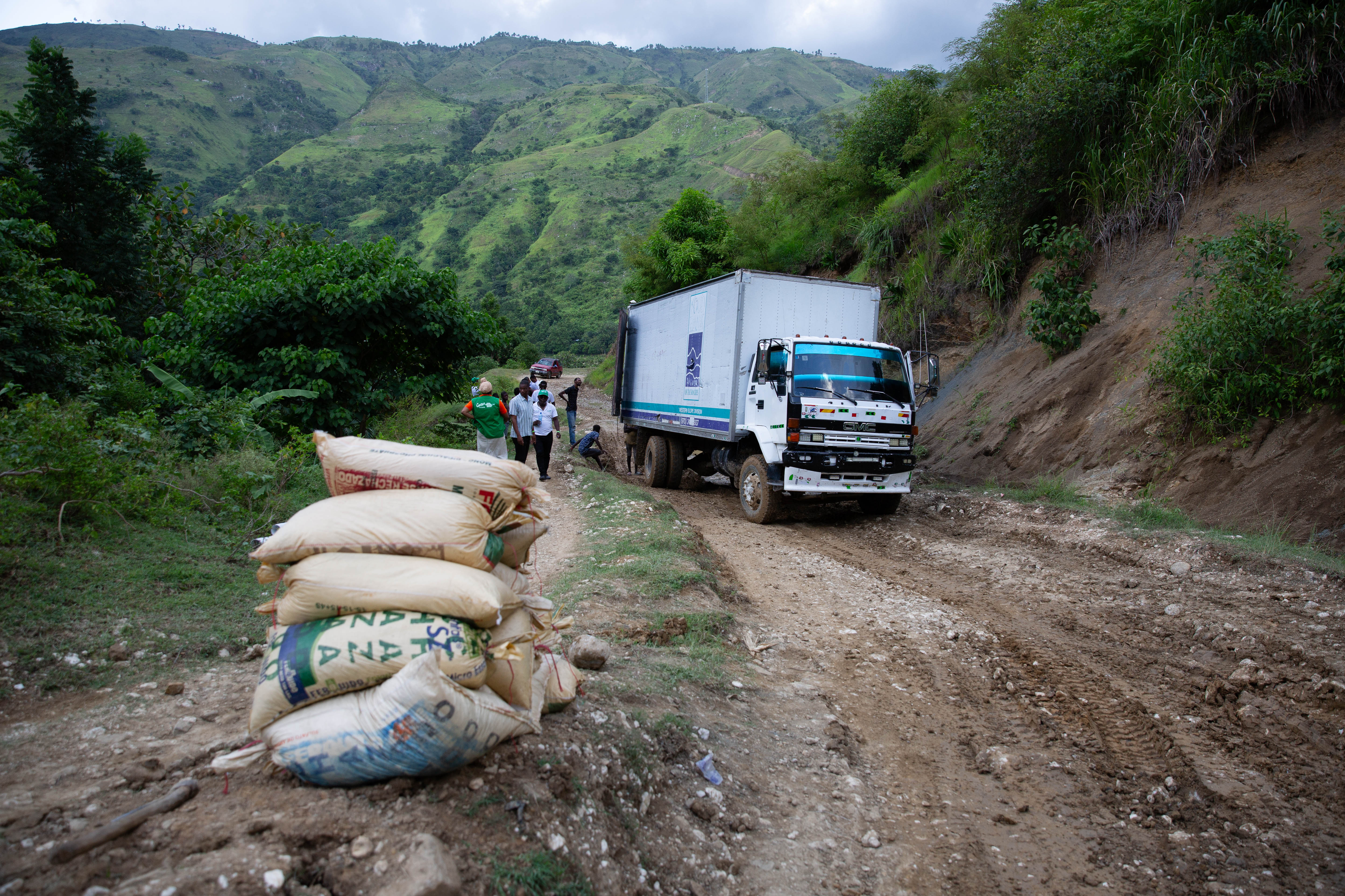 Sacks of supplies piled on the side of a dirt road, with a white truck stopped next to them.