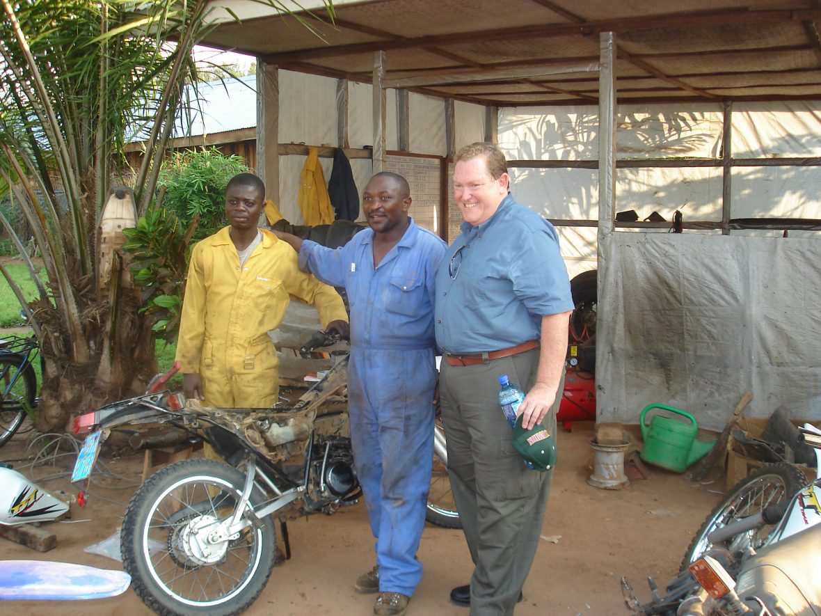 Tom Moran in the DRC posing by two men and a motorbike